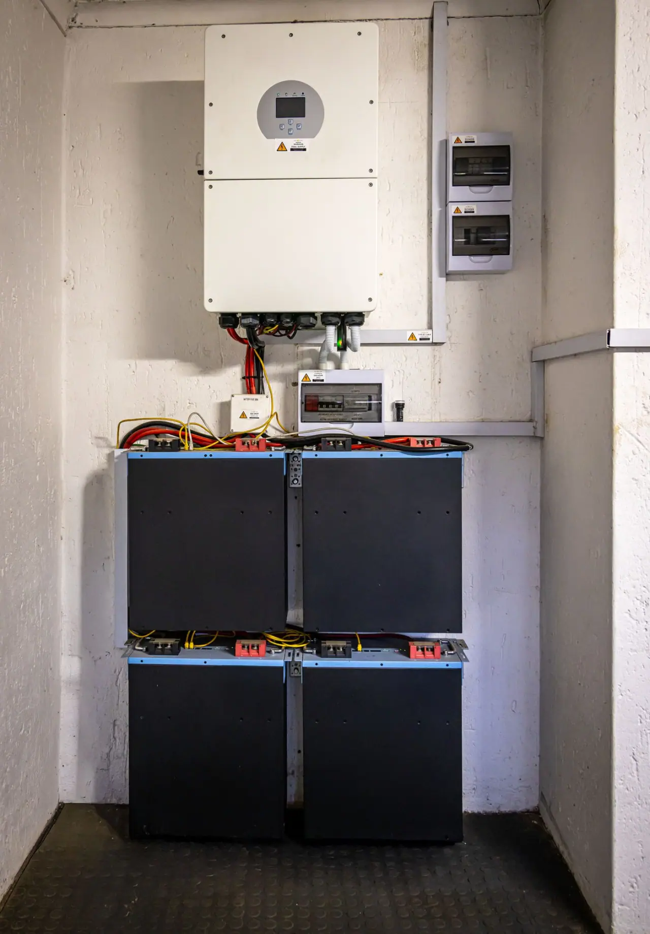 A wall-mounted hybrid inverter with its DC isolator and consumer units, above a bank of four battery modules in a plant room